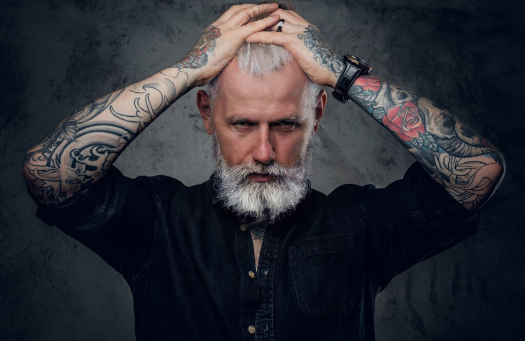 Portrait of old and confident man with tattooed body and gray hairs which poses in dark background.